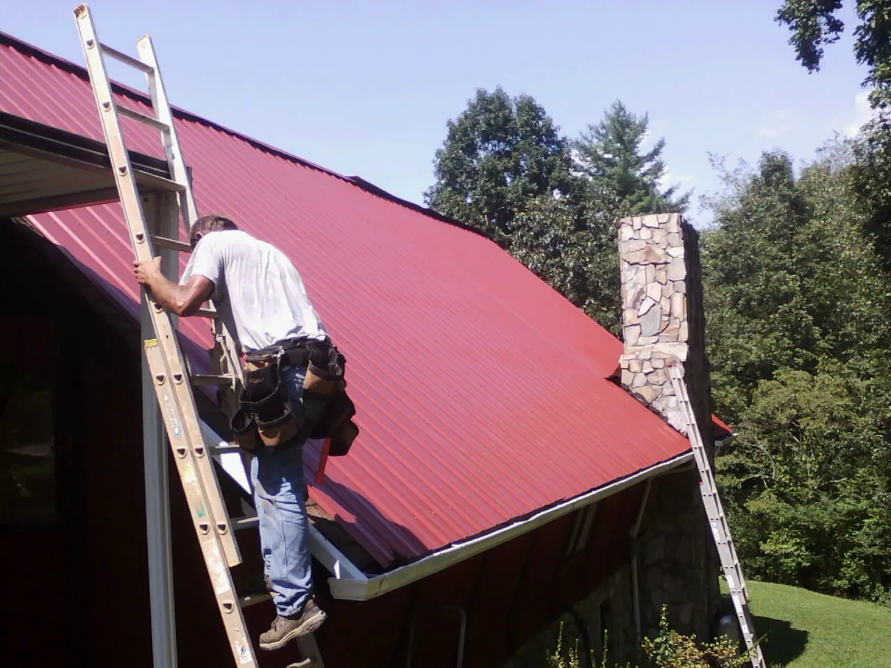 Roofer on a ladder beside a red metal roof and stone chimney — Soffit & Fascia Repair by Roofing Brothers in Tipton